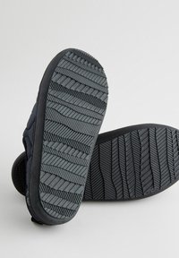 Suole di sneaker in gomma nera con un motivo testurizzato di scanalature e linee angolate. Presentano una forma curva e bordi flessibili per il comfort.