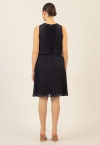 Marineblaues Faltenkleid mit ausgestelltem Rock und ärmellosem Design. Durchsichtiger Stoffüberzug an den Schultern, Reißverschluss auf der Rückseite und weiche Textur.