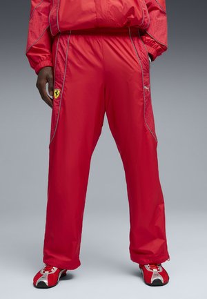 Rode sportbroek met Ferrari- en Puma-logo's, gedragen met bijpassende rood-witte sneakers, vanaf de middel tot aan de voeten getoond tegen een grijze achtergrond.