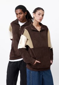 Hnedý zipsový fleece pulóver s béžovými a bielymi rukávmi v blokovanej farbe. Obsahuje vysoký golier a predné vrecká, jemná textúra.