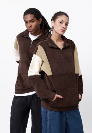 Brown zip-up fleece pullover with beige and white color-blocked sleeves. Features a high collar and front pockets, soft texture.