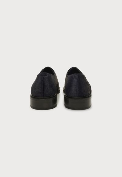 Paire de chaussures habillées noires à petit talon avec dessus texturé, vue de dos sur un fond clair uni.