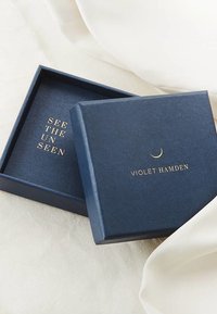 Navyblaue quadratische Box mit strukturiertem Finish. Goldpräge beschrifteter Text lautet "SEE THE UN SEEN" und "VIOLET HAMDEN," ergänzt mit einem Halbmondakzent.