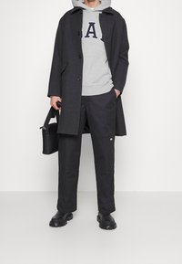 Persona che indossa un cappotto grigio scuro sopra un hoodie grigio GAP, pantaloni neri e stivali neri, mentre tiene una borsa nera davanti a uno sfondo chiaro e semplice.