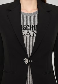 Blazer nero con un design strutturato, caratterizzato da una risvolto pronunciato, un accento in bottone argentato e un maglione metallico testurizzato sotto.