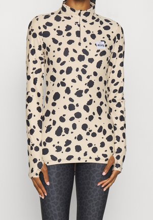 Kobieta ubrana w beżową bluzkę z długim rękawem z czarnymi nieregularnymi plamami i małym naszywką, połączona z ciemnymi legginsami w leopardzi wzór.