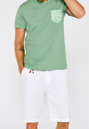 Mann trägt ein hellgrünes T-Shirt mit gemusterter Brusttasche und weiße knielange Shorts, Hand in der Tasche.