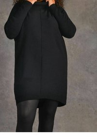 Abito maglione oversize nero con texture a costine, col alto, spalle scese e orlo leggermente curvo. Abbinato a collant neri.