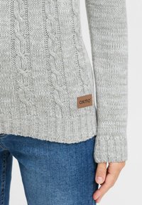 Hellgrauer gestrickter Pullover mit einem Kabeldesign, gerippten Bündchen und einem Lederetikett "oxmo" an der Seite. Weicher, strukturierter Stoff.
