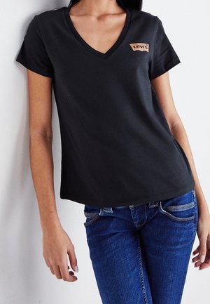 Femme portant un t-shirt Levi's noir à col en V et un jean bleu, debout, les bras détendus, contre un mur blanc.
