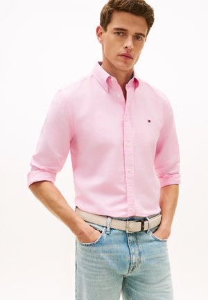 Homme portant une chemise rose clair à boutons avec manches retroussées, un jean bleu clair et une ceinture tressée beige, debout les mains dans les poches.