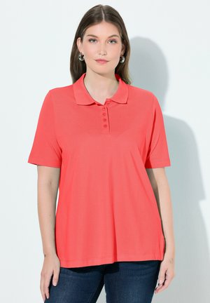 Młoda kobieta z długimi włosami, ubrana w koralową koszulkę polo z krótkim rękawem i ciemne jeansy, stojąca na jasnym tle.