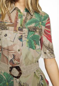 Kurzarmhemd mit einem tropischen Blumenmuster in Grün, Rot und Beige; zwei Fronttaschen und einem strukturierten Gürtel mit einer sechseckigen Schnalle.