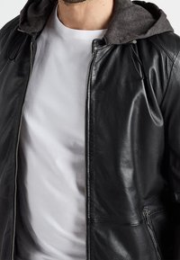 Černá kožená bunda se šedou kapucí. Má stříbrný zip, boční kapsy a hladkou texturu. Nositelná přes bílé tričko.