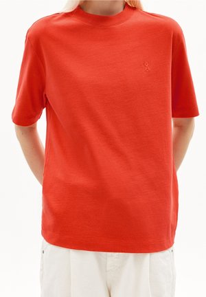 Person trägt ein schlichtes rotes kurzärmeliges T-Shirt mit kleinem geprägtem Logo auf der linken Brust und weiße Hose, Hände hinter dem Rücken.