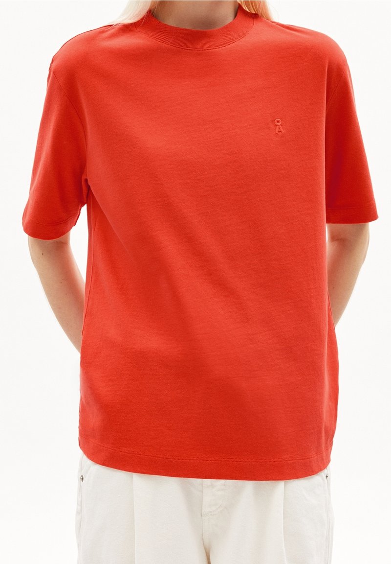Personne portant un t-shirt rouge uni à manches courtes avec un petit logo en relief sur la poitrine gauche et un pantalon blanc, les mains dans le dos.