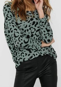 Maglione blu teal chiaro con macchie nere a forma di leopardo, maniche lunghe e vestibilità rilassata, abbinato a pantaloni in pelle nera aderenti. Texture liscia.