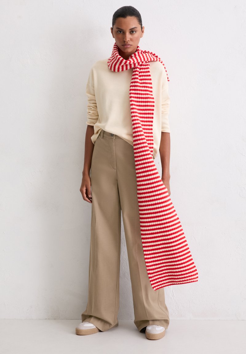 Écharpe à rayures rouges et blanches posée sur un pull clair et un pantalon large beige. Tissu doux et décontracté avec une coupe ample.