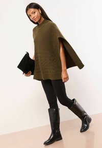 Olivovo zelený pletený pončo s rebrovaným golierom a textúrovaným káblovým vzorom, v kombinácii s čiernymi legínami a čiernymi čižmami po kolená.