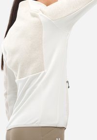 Eine Frau trägt eine crèmefarbene und beige langärmlige Jacke mit Reißverschluss und einem seitlichen Reißverschluss-Taschen. Sie posiert mit erhobenem Arm vor einem einfarbigen Hintergrund.