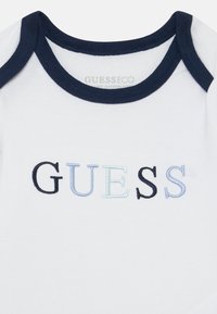 Weißer Baumwoll-Body mit marineblauen Akzenten, versehen mit gesticktem "GUESS"-Logo in verschiedenen Blau- und Schwarztönen. Glatte Textur.