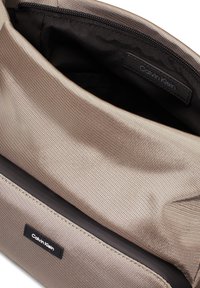 Beige szövet táska texturált külsővel, fekete béléssel és Calvin Klein logóval a belsejében. Zippzáros záródással és elegáns dizájnnal rendelkezik.