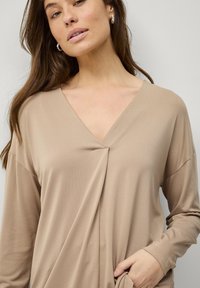 Beige langærmet top med V-hals og draperet for. Glat tekstur med subtil glans, med en løs pasform og sidelommer.