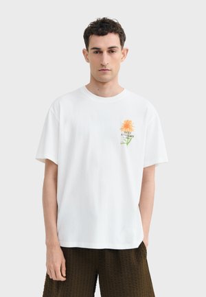 Homme portant un t-shirt blanc avec une fleur orange et le texte "Make a Garden" sur la poitrine, associé à un short texturé foncé, debout devant un fond uni.