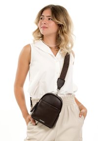 Young woman wearing sleeveless white top and beige pants, carrying small dark brown leather crossbody bag with wide strap.
