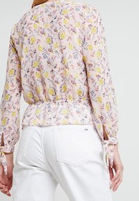 Blusa floreale in tessuto leggero rosa con motivi gialli e blu. Caratterizzata da una vita arricciata e maniche legate. Indossata con pantaloni bianchi.