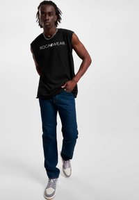 Černé tílko bez rukávů s logem "ROCAWEAR", v kombinaci s tmavě modrými džíny a šedými teniskami. Jednoduchý design s volným střihem.