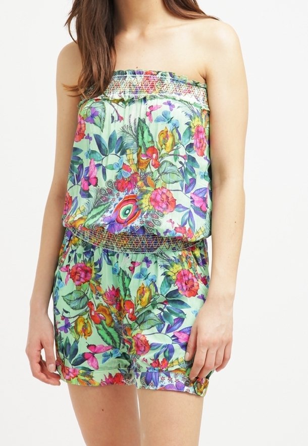 Femme portant une combinaison sans bretelles à fleurs, avec un haut froncé et une taille élastique dans des couleurs vives sur un fond vert clair.