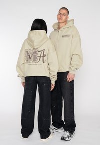 Sweats à capuche beige avec un grand graphique sombre dans le dos, associés à un jean noir à jambes larges. Design décontracté avec des poches bien visibles et des détails de cordon de serrage.