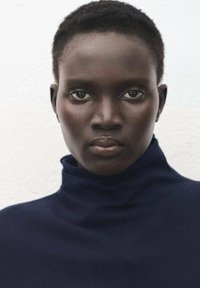 Zbliżenie na osobę w granatowym golfie. Odcień skóry ciemny, o gładkiej fakturze i wyraźnie zarysowanych rysach twarzy. Oczy duże i wyraziste.