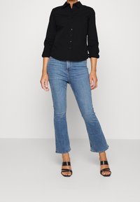 Camisa negra de botones con mangas enrolladas, combinada con jeans de campana azules claros y sandalias negras de tacón, con un diseño minimalista.
