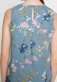 Sininen kukkakuosiset hihattomat puserot, joissa on vaaleanpunaisia, valkoisia ja kultaisia yksityiskohtia. Avainreikätakki yksityiskohta pienen nappisulkimella. Sujuva kangas.