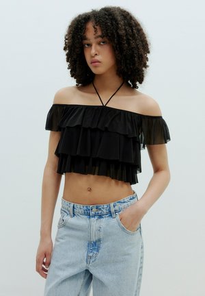 Schwarzes, schulterfreies Crop-Top aus geschichteten Rüschen, mit einer Bindeschließe und kombiniert mit hellblauen, hochtaillierten Jeans.