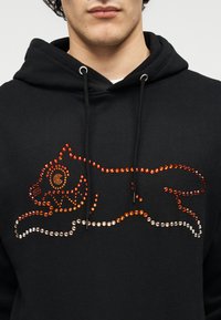 Μαύρο φούτερ με κουκούλα που διαθέτει σχέδιο γάτας διακοσμημένο με πορτοκαλί και ασημί στρας. Κουκούλα με κορδόνια και απαλή υφή υφάσματος.