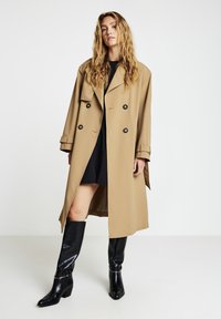 Beiger Trenchcoat mit doppelreihigem Design, schwarzen Knöpfen und einem taillierten Gürtel. Getragen über einem schwarzen Kleid und kombiniert mit kniehohen schwarzen Stiefeln.
