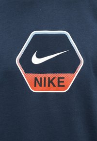 Εξαγωνικό λογότυπο της Nike με λευκό σβήσιμο πάνω από μαύρο κείμενο "NIKE" σε φόντο από ναυτικό μπλε ύφασμα με κόκκινο μοτίβο από τελείες.