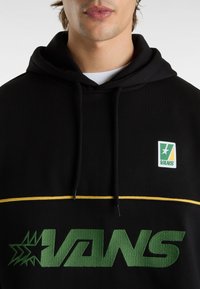 Svart hoodie med grön "VANS"-logotyp, guldkant och liten broderad logotyp på övre vänstra sidan. Material av bomullsblandning med en mjuk textur.