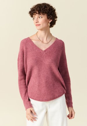 Pull en V, à manches longues, en doux fil rose texturé. Coupe décontractée avec des poignets côtelés, superposé à un pantalon blanc. Détails de design simples.