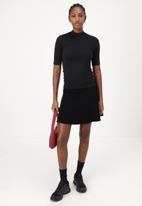 Top nero a maniche corte con collo alto abbinato a una gonna nera a trapezio, indossato con scarpe da ginnastica nere e tenendo una piccola borsa rossa.