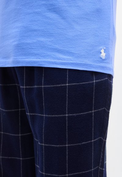 Ανοιχτό μπλε βαμβακερό T-shirt με κοντά μανίκια και μικρό λογότυπο. Συνδυάζεται με παντελόνι πυτζάμας από φανέλα σε σκούρο μπλε καρό με λευκές γραμμές πλέγματος.