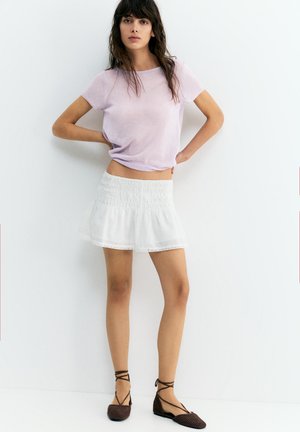 Žena stojaca s rukami na bokoch, oblečená v svetlofialovom tričku s krátkym rukávom, bielej naškrobené mini sukni a hnedých šnurovacích balerínach.