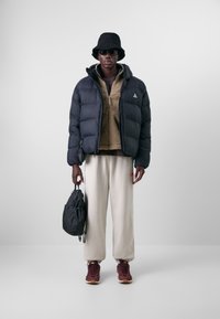 Svart puffjacka, beige fleecetröja med dragkedja, cremefärgade mjukisbyxor, mörk bucket-hatt, solglasögon och vinröda sneakers. Svart väska med axelrem.