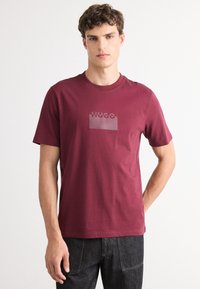 Burgundinės spalvos medvilninė marškinė su apvalia iškirpte, priekyje puikuojasi pusiau permatomas „HUGO“ logotipas blokiniu dizainu.
