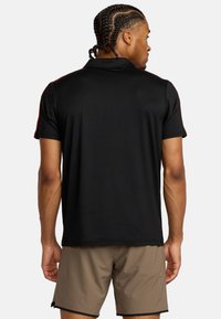 Zwarte poloshirt met korte mouwen, met rode accenten op de schouders, gemaakt van een gladde stof. Draagt een beige korte broek. Achteraanzicht.