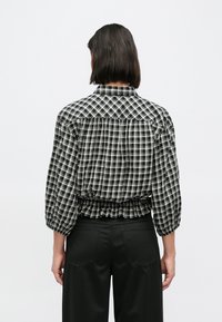 Καρό πουκάμισο σε ασπρόμαυρο με πιέτες στην πλάτη, μανίκια 3/4 και γιακά. Το ύφασμα φαίνεται ελαφρύ και με υφή.