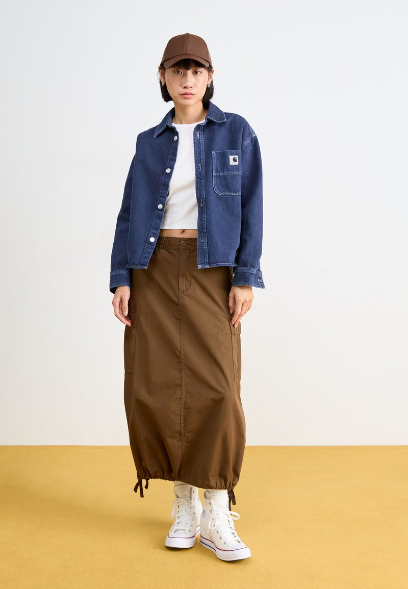Chaqueta de mezclilla en azul oscuro con botones y bolsillo en el pecho, combinada con una falda midi marrón con cordones, usando zapatillas blancas. Gorra marrón.